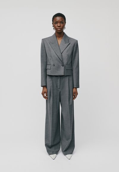 Costume gris sur mesure comprenant un blouson croisé court et un pantalon évasé, complété par des talons aiguilles blancs. Tissu lisse et texturé.