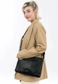 Schwarze, strukturierte Lederhandtasche mit verstellbarem Riemen, rechteckiger Form und metallischen Beschlägen. Über der Schulter getragen vor einem hellen Hintergrund.