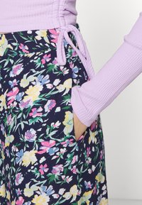 Hand i fickan på en marinblå blommig kjol med rosa, gula och blå blommor, tillsammans med en ribbad lavendelfärgad topp med knytdetalj på sidan.