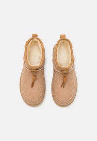 Tan suede ankle boots with a soft, cream fur lining, decorative stitching, and a front tie accent. Features pull tab for easy wear.