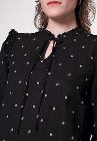 Blusa de tecido preto com riscas verticais, pequenos padrões brancos, ombros com babados e decote em forma de chave com detalhe de laço.