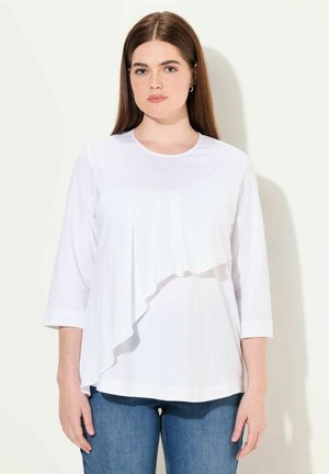 Hvid top med trekvartærmer, der har et lagdelt ruffle-detalje foran. Matchet med medium blå jeans.