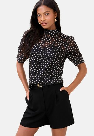 Black polka dot blouse with a high neck, sheer fabric, and puffed sleeves, paired with tailored black shorts and a thin belt.