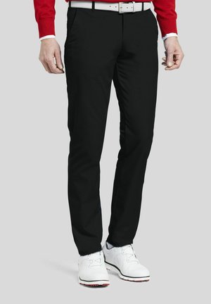 Man draagt strakke zwarte broek, witte riem, rood overhemd met lange mouwen en witte golfschoenen, staand tegen een grijze achtergrond.