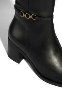 Bottine en cuir noir avec un bout arrondi, un talon bloc de hauteur moyenne, et un accent de chaîne dorée autour de la cheville. Texture lisse et brillante.