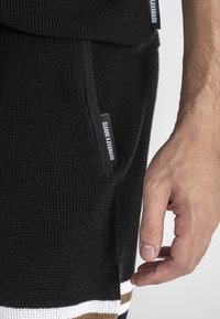 Shorts neri a maglia con strisce orizzontali bianche e marroni all'orlo, dotati di una tasca laterale e un'etichetta con la scritta "GIANNU KAVANAGH."