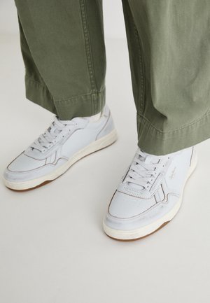 Baskets blanches en cuir avec semelles beige portées avec un pantalon vert olive, debout sur un sol clair.