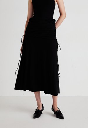 Abito nero con vita aderente e texture arricciata, caratterizzato da lacci laterali. Abbinato a scarpe nere a punta su uno sfondo chiaro.