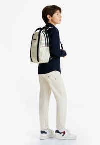 Gestreepte rugzak in roomwit en marineblauw, met logo en zwarte accenten. Draag op een kind in lichtbeige broek en witte sneakers.