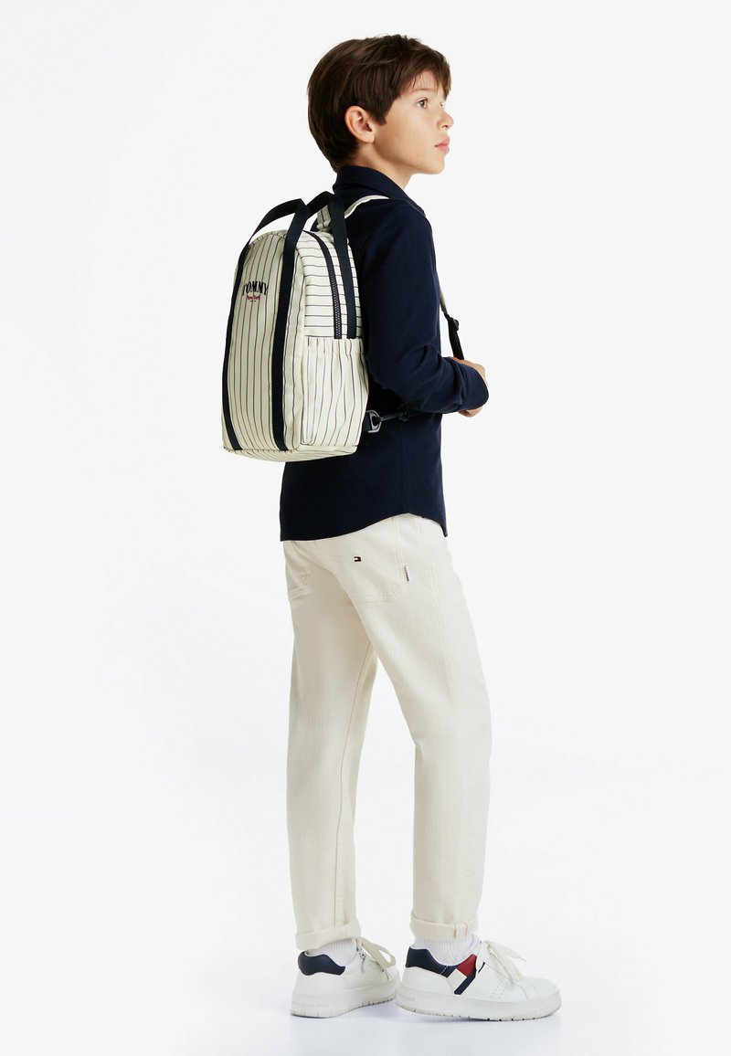 Gestreepte rugzak in roomwit en marineblauw, met logo en zwarte accenten. Draag op een kind in lichtbeige broek en witte sneakers.