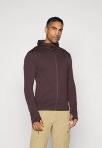 Bordo zip-up kaput s uskim dizajnom, mekim materijalom, visokim ovratnikom i rukavima s otvorom za palčeve. Nositi preko svijetlosmeđih hlača.