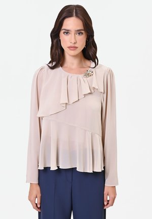 Donna con i capelli scuri indossa una camicetta beige chiaro con volant e spilla dorata, abbinata a pantaloni blu navy, in piedi su uno sfondo bianco.