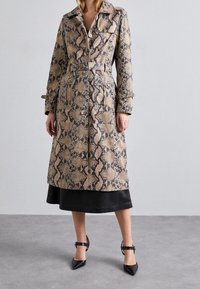 Trench-coat Python beige et noir, avec une taille ceinturée, un col pointu et des fermetures à boutons. Porté sur une jupe noire.