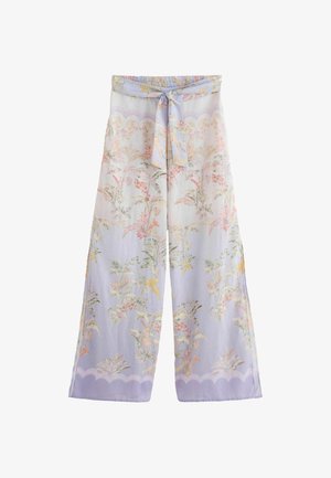 Kobieta stojąca boso w kwieciste, szerokie spodnie i dopasowaną bluzkę z długim rękawem zawiązaną w talii na tle kamiennej ściany.