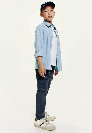 Camicia button-up azzurro chiaro sopra una maglietta bianca, pantaloni cargo scuri e sneakers bianche con dettagli blu navy. Il cappellino è blu navy con un logo.