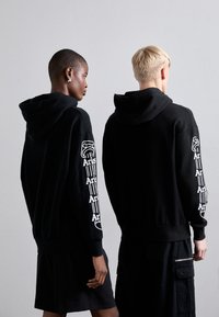Zwarte hoodies met witte bedrukte tekst en graphics op de mouwen. Katoenen stof, losse pasvorm, met ribgebreide manchetten en onderrand.