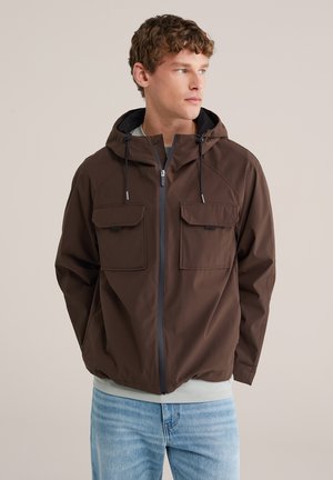 Man met krullend haar die een bruine capuchonjas met voorzakken en lichtblauwe spijkerbroek draagt, kijkend naar rechts tegen een effen achtergrond.