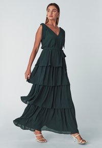 Dunkelgrünes, ärmelloses Maxikleid mit V-Ausschnitt, gestuften Lagen und einem Stoffgürtel. Leichte Textur mit subtilen Drapierungen und einer lässigen Silhouette.