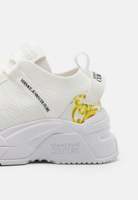 Baskets blanches slip-on avec semelle épaisse, motif baroque jaune au talon et marquage "Versace Jeans Couture" sur le côté et la semelle.