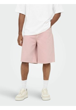 Osoba nosiaca svetloružové krátke nohavice pod kolená, biele tričko, biele podkolienky a krémové tenisky, stojaca s jednou rukou v vrecku.