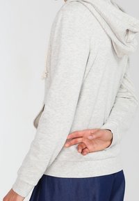 Sudadera gris claro con un bolsillo canguro, cordones ajustables y puños acanalados. Textura suave y corte relajado. Pantalones azules visibles por debajo.