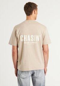 Beige bomulds t-shirt med hvidt fed skrift "CHASIN'" og mindre tekst nedenunder, korte ærmer og en afslappet pasform.