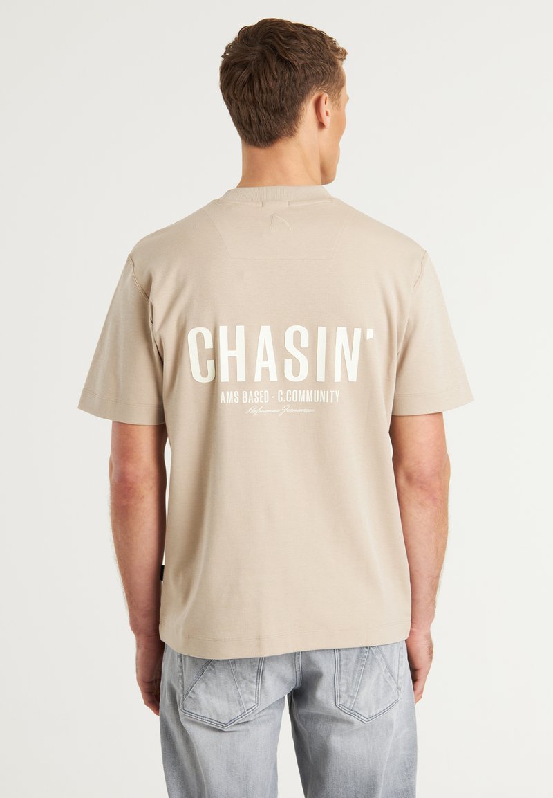 Beige bomulds t-shirt med hvidt fed skrift "CHASIN'" og mindre tekst nedenunder, korte ærmer og en afslappet pasform.