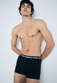 Boxer shorts noirs en tissu doux avec une large ceinture noire arborant le logo blanc "KARL LAGERFELD". Modèle aux cheveux bouclés.