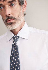 Camicia bianca con colletto rigido, abbinata a una cravatta blu scuro con pois crema. Tessuto liscio e vestibilità su misura.