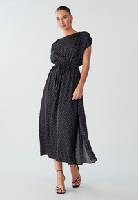 Schwarz gepunktetes Maxikleid mit runder Ausschnitt, elastischem Bund, Seitenschlitz und kurzen Ärmeln. Fließender Stoff mit einer glatten Textur.