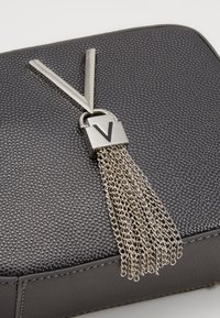 Materiale nero testurizzato con un accentuato hardware argentato, caratterizzato da un lucchetto e una frangia a catena. Il lucchetto presenta un logo "V" ben visibile.