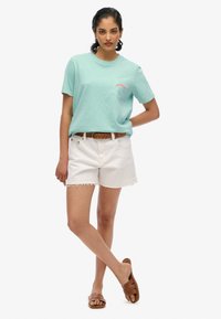 Lysblå kortærmet t-shirt med en lomme, med rød tekst. Sammen med hvide denimshorts og brune slidesandaler.