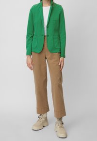 Zelený bavlněný blazer se dvěma předními kapsami a klopami, kombinovaný s béžovými kalhotami a hnědými kotníkovými botami.