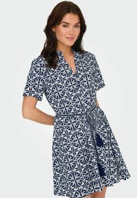 Kratka haljina s kratkim rukavima u bijeloj i mornarskoj plavoj boji s geometrijskim uzorkom, ovratnikom i vezicom s resicama u struku.