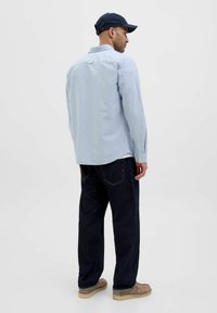 Camicia azzurra chiara a maniche lunghe con dettagli sul retro, abbinata a jeans blu scuri e scarpe beige. Indossata con un cappellino blu navy.