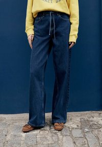 Jeans de mezclilla azul oscuro de pierna ancha con cintura alta, con un lazo frontal y costuras visibles, combinados con zapatos estilo mocasín marrón.