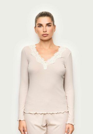 Mujer con el cabello recogido, lleva una camiseta de manga larga color beige claro con detalle de encaje en el escote en V y pantalones a juego, mirando hacia adelante.