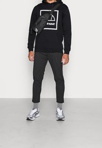 Czarna bluza z kapturem z graficznym logo, czarne spodnie w prążki, szare skarpetki i srebrne buty sportowe. Czarna torba na ramię noszona na krzyż.