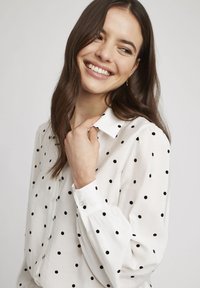 Blouse blanche à pois noirs, boutonnière sur le devant, col classique, et manches longues avec poignets froncés. Texture de tissu lisse.