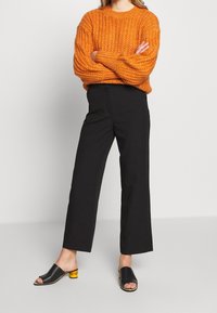 Oranje gebreide trui met een gestructureerd patroon, gecombineerd met hoge zwarte wijde broek en zwarte instapsandalen met een blokhak.