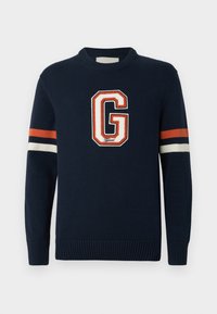 Tamsiai mėlynas megztinis, turintis didelį baltą ir oranžinį "G" logotipą, o ant rankovių matomos oranžinės, baltos ir tamsiai mėlynos spalvų juostos. Raukšlėti rankogaliai.