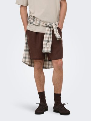 Persoon die een beige T-shirt draagt, bruine korte broek, donkerbruine sokken en schoenen, met een beige en bruine geruite blouse om de taille gebonden.