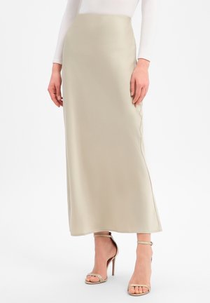 Frau trägt einen beigen, knöchellangen Rock, kombiniert mit hautfarbenen Sandalen mit hohen Absätzen und einem weißen Langarmshirt vor einfachem Hintergrund.