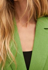 Blazer vert en tissu texturé, avec des revers crantés et un col ouvert, porté sur un haut noir. Les cheveux tombent en ondulations douces autour du cou.