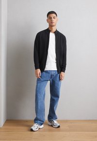 Svart hettejakke med glidelås over en hvit T-skjorte, kombinert med lyseblå vide jeans og grå joggesko. Glatt stoff og avslappet passform.