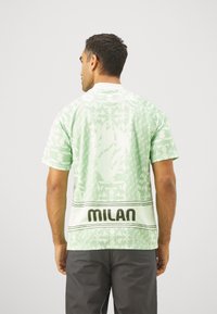 Camisa de manga corta verde menta con patrones geométricos y un gran logo de "MILAN" en el dobladillo. Hecha de un tejido ligero.