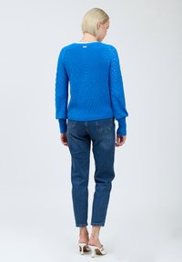 Blauer gestrickter Pullover mit strukturierten Ärmeln, geripptem Saum und Rundhalsausschnitt. Kombiniert mit dunklen Denim-Jeans und weißen hohen Sandalen.