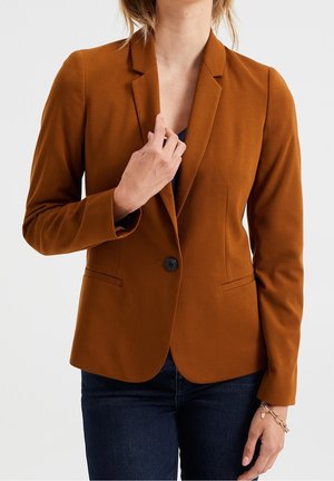 Vrouw draagt een aangesloten roestkleurig blazer met één vastgeknoopte knoop, gecombineerd met donkere jeans, tegen een eenvoudige witte achtergrond.