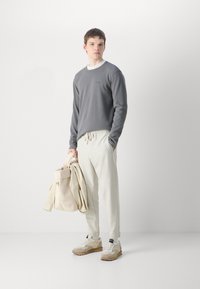Chemise grise à manches longues avec texture côtelée, pantalon crème avec cordon de serrage et baskets beiges. Tient un sac beige clair. Look simple et moderne.