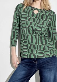 Maglietta a maniche lunghe verde con motivo e grandi numeri neri, con un dettaglio a chiave sullo scollo. Abbinata a jeans blu scuro.